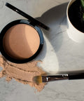 Compact powder with a brush on a marble surface next to a small plant
