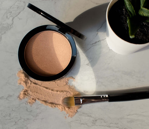 Compact powder with a brush on a marble surface next to a small plant
