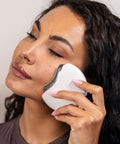 Woman using a white facial cleansing brush on her face against a neutral background
