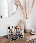 Clear acrylic cosmetic organizer on a wooden surface with makeup items, a mirror, and decorative elements.